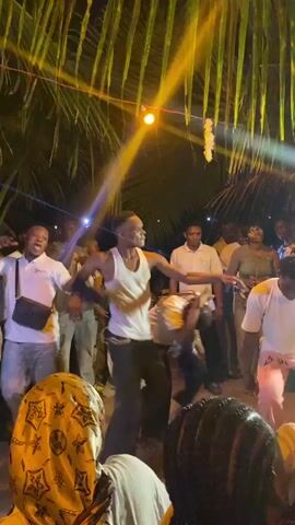 The video captures a lively scene of people dancing at an outdoor gathering at night. The crowd is diverse, with individuals dressed in various casual outfits. The lighting is a mix of yellow string lights, white spotlights, and the flash from phone cameras. The atmosphere appears energetic and celebratory. Palm fronds hanging overhead suggest a tropical or beach-like location. Some attendees are holding their phones up to record the dancing.