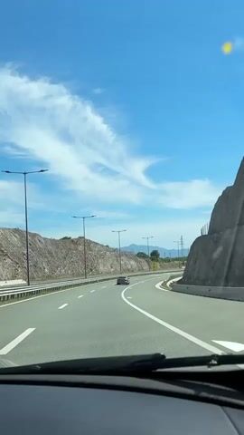 This is a first-person perspective video shot from inside a car driving on a highway. The road is paved and has multiple lanes with clear white markings. There are several streetlights lining the highway. On both sides of the road, there are rocky hills with sparse vegetation. The sky is bright blue with some wispy white clouds. In the distance, mountains are visible. There are a few other cars on the road, all moving in the same direction. Some speed limit signs (80 km/h) are visible on the side of the road. The car's windshield wipers are visible at the bottom of the frame. The overall scene suggests a sunny day with good visibility on a highway in a hilly or mountainous region.
