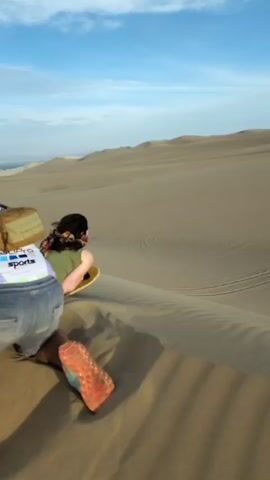 A woman sandboards down a sand dune while lying on her stomach on a green and yellow board. Another person can be seen in the background walking further down the dune. The first woman wears a green shirt and denim shorts. A GoPro sports shirt is visible in the foreground. The sky is blue with white clouds.