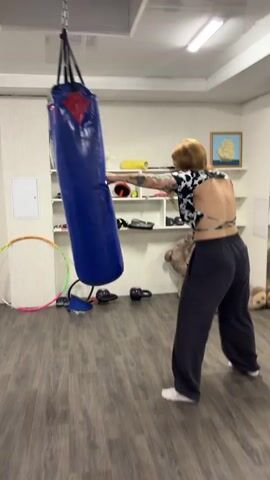 A woman with blonde hair and tattoos is punching a blue punching bag in a room with a wooden floor. She is wearing a black and white patterned top and dark pants. In the background, there are shelves with various items, including a teddy bear and a hula hoop. The video then cuts to a different scene showing a woman with glasses and brown hair tied up, wearing pajamas, jumping rope over a blue ping pong table. The lighting is bright and appears to be indoors.