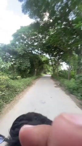 The video shows a first-person perspective of someone riding a motorcycle on a paved road surrounded by lush green trees. The trees form a canopy over the road, creating a tunnel-like effect. The sun is shining, and the sky is visible through the leaves. The rider's head and shoulders are visible in the foreground, indicating the camera is mounted on their helmet or is being held by them. The road appears to be in a rural area, with vegetation on either side. The rider is moving at a moderate speed, as indicated by the slight blur of the trees. The video is short, lasting only a few seconds, and captures a peaceful and scenic moment of travel.