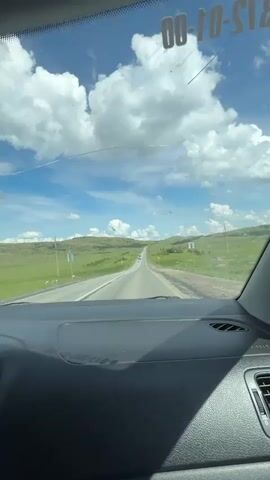 The video captures a view from inside a car looking out through the windshield. The road ahead is straight and stretches into the distance, bordered by rolling green hills. The sky is blue with fluffy white clouds. Several vehicles are visible on the road, including cars and a white bus with writing on its side. The dashboard of the car is partially visible in the foreground. The car appears to be moving along the road.