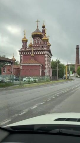 The video captures a view from inside a vehicle driving on a wet road. To the left, a red brick church with golden domes is visible, bordered by a fence and some greenery. Further down the road, industrial structures with tall chimneys dominate the landscape, suggesting a factory or industrial complex. The sky is overcast and grey. The vehicle appears to be moving forward, with the road curving slightly to the right. A speed bump is visible on the road as the vehicle approaches it. Several cars are seen on the road, and a pedestrian crossing is also present. The overall atmosphere is industrial and somewhat bleak, with the church offering a contrasting element.