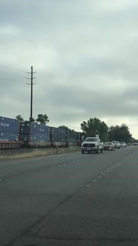 This media file captures a scene on a road in Roseville, United States of America, under an overcast sky, suggesting a cloudy or potentially rainy day. The perspective is from inside a vehicle, looking forward.

The primary subjects are vehicles on a multi-lane road. In the foreground, a white Ford pickup truck is prominently visible, followed by a red sedan, a white SUV, and a black sedan. Further down the road, other vehicles, including trucks and cars, can be seen. To the left of the road, stacked shipping containers are visible, some with blue and white markings. Beyond the containers and across the road, lush green trees line the landscape. Power lines and utility poles are also present, indicating infrastructure in the area.

The activity taking place is traffic flow on a public road. The vehicles appear to be in motion, suggesting a commute or travel. There are no people clearly visible within the vehicles.

Notable details include the time of day, which is difficult to ascertain precisely due to the overcast conditions, but the lighting suggests it is daytime. The road itself has yellow reflectors marking the lanes and some visible cracks in the asphalt, indicating wear and tear. The presence of shipping containers might suggest proximity to a port, railyard, or distribution center.

No significant text is clearly legible on the vehicles or the shipping containers, though some branding might be present on the white pickup truck. The overall mood is neutral, focusing on the mundane aspect of road travel.