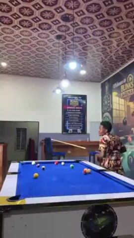 Here is a concise description of the video:

A young man stands near a pool table with a blue felt surface in what appears to be a billiards hall. He is holding a pool cue and seems to be reacting with enthusiasm, possibly after a shot. The pool table has several pool balls on it, and the environment includes overhead lighting, chairs, and signage related to "Blinks Arena," which seems to be the name of the billiards hall. The decor features a patterned ceiling and a mural with billiard-related imagery on the wall.