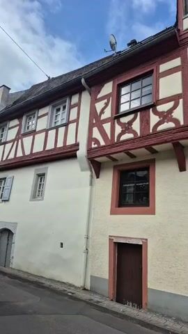 A low-angle shot captures a historical building in Germany, with white and red walls, and dark wooden window frames and doors, on a partially cloudy day. The building, a traditional German house, has a red half-timbered upper facade with a light-colored infill. Two dark-framed windows are visible on the first floor, and a dormer window extends from the roof. A small brown door with a semicircular arch is at the bottom right. The building is attached to another one on the left, which has a white facade and a single window on its lower level. The sky above is blue with some white clouds.
