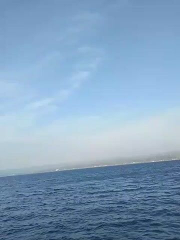 The video shows a wide shot of the ocean under a pale blue sky with some wispy clouds. The water is choppy and a deep blue. In the distance, a hazy shoreline is visible with a few faint white specks that could be buildings or boats. The overall impression is of being out on the water on a somewhat overcast day. The camera appears to be moving slightly, suggesting it's being held by someone on a boat. There are no people or specific objects clearly identifiable in the frame.