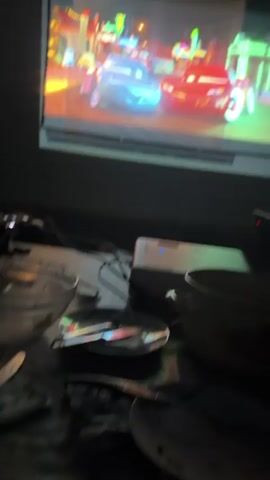 The video shows a projector screen displaying a scene from the animated movie "Cars." Two cartoon cars, a blue female car and a red male car, are side-by-side in what appears to be a town at night, with neon signs in the background. Below the screen, a projector is visible, along with various electronic devices and cables. In the foreground, there are several bowls and other unspecified items. The room is dark, with the primary light source coming from the projector.