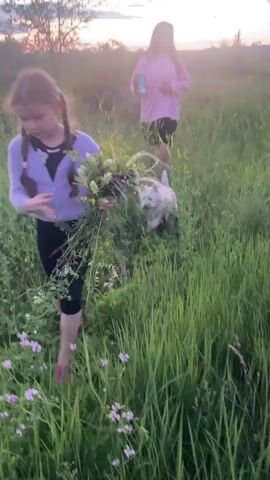 The video shows two young females and a white dog in a grassy field with flowers. The female in the foreground is wearing a purple long-sleeved shirt and black leggings, bending down to pick flowers and hold a bunch of greenery. She has dark hair in two braids. The female in the background is wearing a pink long-sleeved shirt and black shorts, walking away from the camera. The white dog, a West Highland White Terrier, is in the foreground, near the female who is picking flowers. The sun is setting in the background, casting a warm glow. The environment is a natural, outdoor setting with tall grass and scattered flowers. The video appears to capture a moment of leisure and exploration.