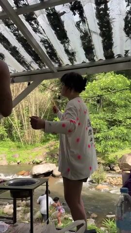 A woman wearing a white long-sleeved shirt with pink polka dots stands outdoors near a river. She is holding chopsticks in her hands and appears to be talking. In the background, a man is holding a baby by the river, and there is a grill on the left side of the frame. Bamboo trees and lush greenery surround the river.