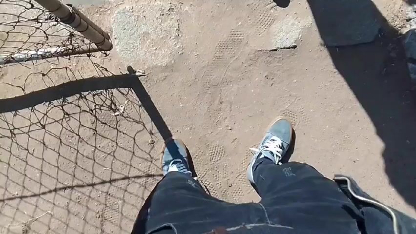 The video features two individuals outdoors in Klerksdorp, South Africa, on a bright, sunny day, indicated by strong shadows. The primary subject is wearing a black furry bucket hat, a black long-sleeved shirt, a black zippered vest, black ripped jeans, a brown belt, and light blue/grey sneakers with white laces. They also have a silver cross necklace.

The activity involves the primary subject posing for the camera, adjusting their hat and clothing, and showcasing their outfit. At one point, they bend down and then stand back up, continuing to pose. Towards the end of the video, a second person briefly appears, and the primary subject engages in a fist-bump with them. The second person's feet are visible, wearing red sneakers and black track pants with white stripes (identifiable as Adidas).

The scene is set on sandy ground next to a wire fence, with a paved area visible. In the background, there are trees, some bare and some green, and what appear to be residential buildings under a clear blue sky. No visible text is present in the video frames.