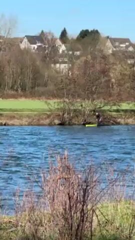 The video depicts a person standing on a yellow and black paddleboard in a body of water. The person is wearing a dark wetsuit with white stripes on the arms and is holding a paddle. They appear to be pushing off from the edge of the water near some vegetation and trees. The water is choppy with visible waves. In the background, there are houses and more trees under a clear blue sky. The scene appears to be set in a natural or semi-natural environment.