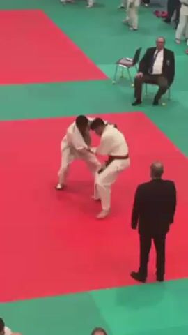 Two judoka wearing white gi suits compete on a judo mat which is red and green. One judoka wears a red belt, the other wears a green belt. The judoka in the red belt throws the judoka in the green belt to the ground. An official in a black suit walks towards the competitors. Another man in a black suit is sitting on a chair in the background. Other people in judo uniforms can be seen in the background.