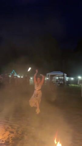 A woman with blonde hair tied back is dancing in a sandy, outdoor area at night. She is wearing a light-colored dress with ruffled trim on the sleeves and hem, and white boots. She is holding a flaming prop in her right hand, which she is twirling around her head. She appears to be performing for a crowd of people in the background, some of whom are illuminated by string lights. There are trees and tents visible in the background, and the ground is covered in sand and dirt. Smoke or fog is swirling around the dancer.