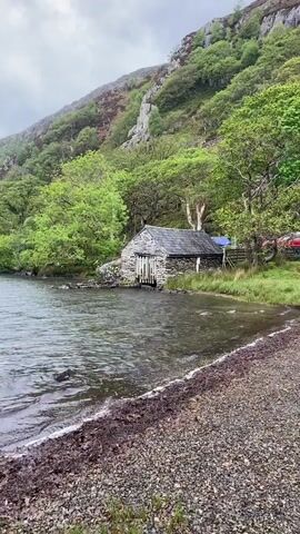 The video shows a lake scene on a cloudy day. The lake is surrounded by green hills and trees. There is a stone building with a gray slate roof on the shore. A person in a black jacket and gray pants walks to the shore and throws a rock into the water. The camera then pans across the lake to show the wider view of the surrounding hills and cloudy sky. There are a few birds flying in the distance. The water is choppy and gray, and the shoreline is made of small pebbles.