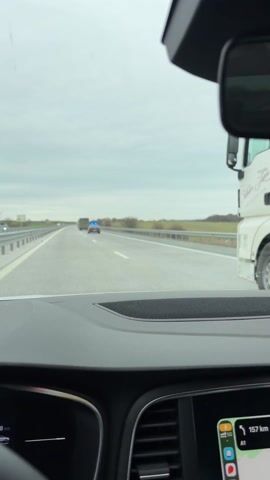 The video shows a vehicle traveling on a highway behind a "Military Police" car with flashing blue lights and two olive-colored buses. The Military Police car is a black sedan with the words "Military Police" printed on the back. The buses are in front of the car, and the Military Police car is directly behind the first bus, with a second truck in the distance. The highway is gray asphalt with white lane markers, and there is a green grassy area with trees visible to the right of the highway. The sky is overcast and gray. The view is from inside a car, looking out over the dashboard.