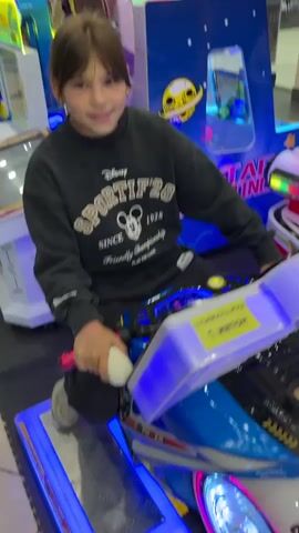 A young person with dark hair is sitting on a blue and white arcade motorcycle game. They are wearing a black sweatshirt with white writing on it. The motorcycle has flashing pink and blue lights on its wheels and handlebars. The game has a yellow sign on it that says "стоимость игры 1 жетон" which translates to "game cost 1 token". The person is holding onto the handlebars and smiling at the camera. In the background, there are other arcade games and people walking around in what appears to be a mall. The floor is tiled.