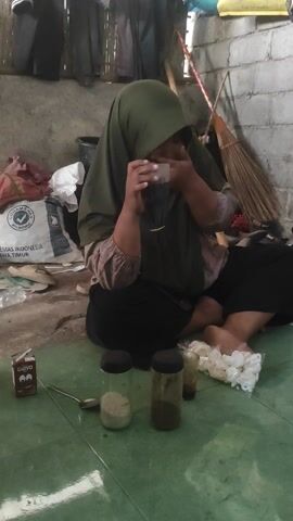 A person wearing a green hijab and a brown shirt is sitting on the floor, facing left. Their head is bowed, and their hands are covering their face. To the right of the person is another person wearing a black outfit and black hijab, also sitting on the floor. Between them and further to the right are several objects including a broom, a bag of white items, two glass jars with dark lids, a small white cylindrical object, and a small dark box. The floor appears to be a mix of concrete and green tiles. The environment seems to be an indoor space with a rough concrete wall in the background and some clothing items hanging on a rack.