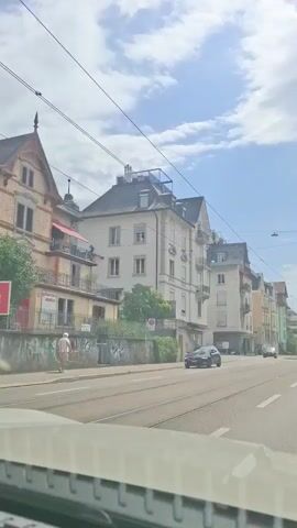 The video shows a street view from a vehicle. Buildings line the right side of the street, some with balconies and awnings. Overhead power lines are visible. A black car is driving in the left lane of the road, and a person appears to be hanging from a wire between two buildings on the left side of the street. A white railing is visible in the foreground, suggesting the footage is from inside a car. The sky is partly cloudy with blue sky visible.