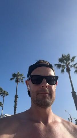 The video is a first-person selfie taken outdoors on a sunny day with a clear blue sky. The subject, a man wearing sunglasses, a baseball cap, and earbuds, is shirtless. He is standing with palm trees and a white building in the background. At 00:01, the man raises his right index finger and then his thumb and index finger together as if to indicate a small size. At 00:03 and 00:04, a woman and a dog are visible in the background walking on a paved path. The man appears to be at a beach or a coastal area.