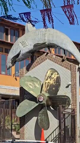 This is a close-up shot of a large metal shark sculpture mounted above a large metal ship propeller. The shark is gray and appears to be made of overlapping metal plates. It has its mouth open, revealing rows of sharp teeth. The propeller is green and brass-colored with four blades and is mounted on a wall decorated with a brown and tan tile pattern. Below the propeller, an orange traffic cone is visible. In the background, there is a building with orange and brown walls and windows. Colorful pennants or streamers are strung across the top of the frame. The lighting suggests it is daytime with the sun casting shadows.