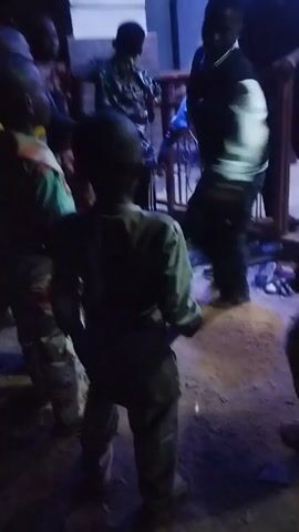 The video shows a group of people gathered outside a building at night. The setting appears to be a residential area, with a tiled building and metal gate visible. The group mainly consists of children, who seem to be moving around. There is at least one adult standing near the gate. The lighting is dim, with some areas more illuminated than others, casting shadows and making details difficult to discern. The overall impression is a casual gathering in a dimly lit environment.