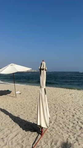 The video shows a sunny beach scene with blue skies. Two beach umbrellas are visible. One is open, casting a shadow on the sand, while the other is closed and standing upright. In the background, the ocean meets the shoreline with gentle waves. A faint outline of a town or settlement is visible in the distance. The sand appears clean and undisturbed.