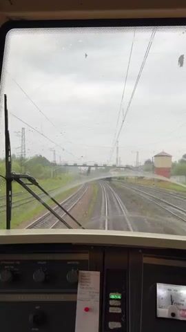The video is filmed from the perspective of someone in the driver's cabin of a train. The windshield is wet with raindrops, and a windshield wiper is visible on the left side. Through the windshield, multiple sets of train tracks stretch out ahead, curving to the right. A train with visible cargo cars is moving along one of these tracks. Overhead, electrical catenary lines are present, supported by poles. In the distance, there are some trees, industrial structures, and a water tower. The dashboard of the train cabin is partially visible at the bottom of the frame, with various buttons, dials, and a gauge with a needle. The sky appears overcast and cloudy.