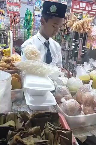 A young boy wearing a white shirt, a tie, and a black peci hat with an emblem is standing behind a market stall. He is looking down at a stack of white foam containers and a pile of white crackers. The stall is filled with various food items packaged in plastic bags and containers. In the foreground, there are several food items wrapped in banana leaves. The background shows shelves stocked with packaged goods. The lighting appears to be artificial, suggesting an indoor market setting. The boy's expression is focused on the task at hand.
