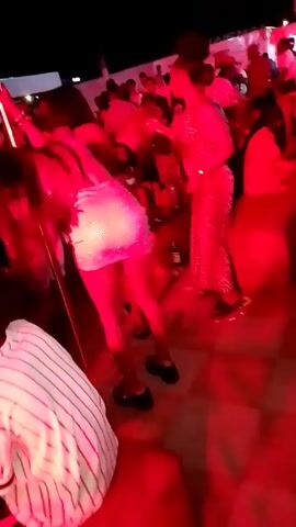 The video shows a crowded outdoor party at night with red lighting.  Several people are dancing. A woman in a shiny mini-dress dances near a pole, bending over and moving her hips. To her right is a woman in a leopard-print dress. The foreground includes a person in a striped shirt.  Other partygoers can be seen in the background.