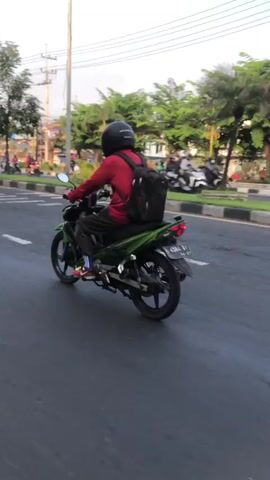 The video shows a person riding a green motorcycle on a paved road. The rider is wearing a red shirt, black pants, a black helmet, and a black backpack. The motorcycle has a license plate. The road has lane markings and is bordered by trees and other vegetation. In the background, other vehicles and people can be seen. The lighting suggests it is daytime.