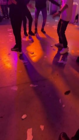 The video shows a group of men dancing at what appears to be a celebration or party. The lighting is colorful, shifting between blue, white, and pink. Confetti-like scraps are scattered on the floor. One man stands on a chair while others dance around him, raising their arms. The men are dressed in casual clothing, including jeans, t-shirts, and button-down shirts. The room has a low ceiling covered with white fabric. Balloons and other decorative elements are visible in the background.