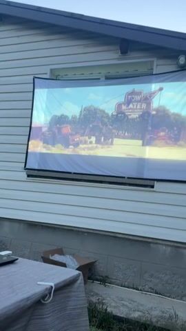 The video shows a projection of the animated movie Cars on a screen mounted on the side of a house. The scene on the screen features the character Mater and a sign that says "TOW MATER". The house has white siding and a window behind the screen. In the foreground, a table covered with a gray tablecloth is visible. There is also a cardboard box on the ground near the table and some green grass. The sky appears clear and blue.