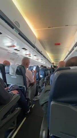 The interior of an aircraft is shown. Passengers are seated, and one person is using their phone. A flight attendant wearing a green uniform is standing in the aisle, presumably speaking to the passengers. There are overhead lights on, and signs indicate exit locations. The seats are blue, and the aisle is narrow.