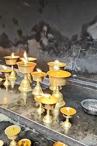 A video pans across a table filled with lit brass oil lamps. The lamps are of various sizes and designs, some with decorative bases. The flames flicker, casting a warm glow on the surrounding area. The table appears to be made of a dark, reflective material, possibly marble or polished stone, and is slightly wet, with some drips of what looks like melted wax. Behind the table is a dark, textured wall. The overall impression is of a dimly lit, devotional space.