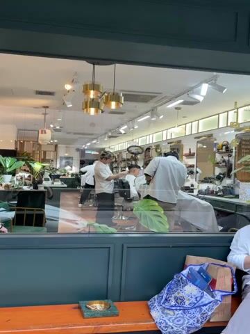 The video shows the interior of a barber shop, as seen through a window. Inside the shop, several barbers are working on customers. Two barbers are actively cutting hair. The shop is well-lit with multiple overhead lights and hanging fixtures. The decor includes mirrors, plants, and barbering tools displayed on shelves. Outside the window, a wooden bench is visible, along with a patterned bag and a small green tray. The window frame is painted a dark green.