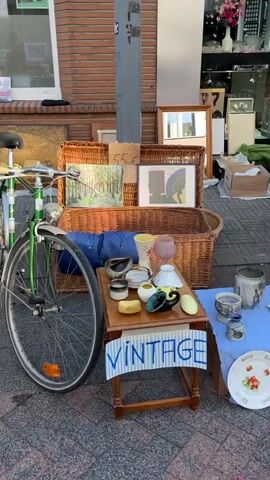 The video pans across a street sale. In the foreground, a green bicycle is parked. To the right, a small table displays various ceramic items, including a vase and bowls, and a sign that reads "VINTAGE". Further back, two chairs are set up, one with a framed picture and the other empty. Behind the chairs, a large basket is placed, and next to it, framed artwork and other items are displayed. On the left, a large framed painting is leaning against a brick wall. More items, including boxes and what appears to be a sculpture of a detached head, are visible. In the background, a building with windows and a door is visible, with tables set up in front of it, displaying more goods, including books, and what looks like more artwork. The sale appears to be taking place outdoors on a paved surface. A person walks by in the foreground of the final shot.