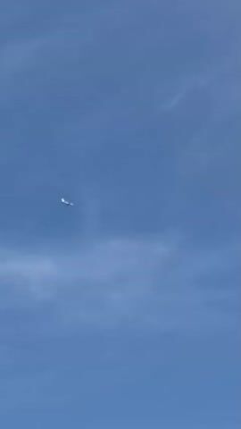 A video recording shows a bright blue sky with a few wispy white clouds. A small, white object is visible in the upper left quadrant of the frame, appearing to be high in the atmosphere. The object is angled, with one end appearing slightly wider than the other, suggesting it may be an aircraft. There are no other objects or people visible in the frame.