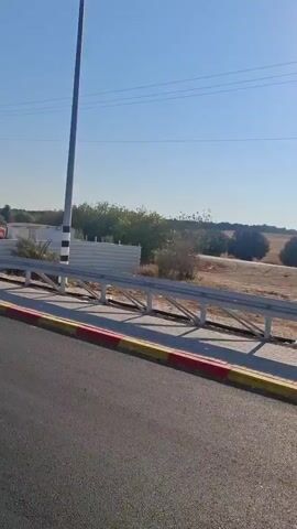 The video shows a roadside view from a moving vehicle. The road has yellow and red painted curbs.  To the right, a black and white striped pole, likely a light pole or utility pole, stands tall.  Beyond the road, a temporary barrier made of orange plastic cubes is visible.  Further in the background, there are construction materials, sand piles, trees, and a few structures that appear to be portable buildings.  Power lines are visible overhead. The sky is clear and blue, indicating a daytime scene.
