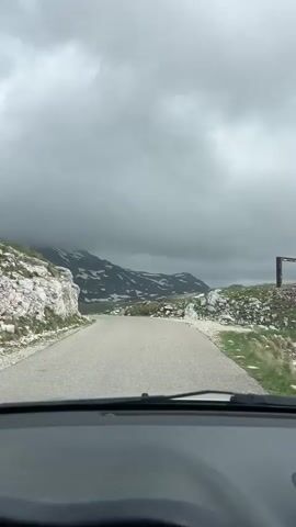 The video captures a first-person view from inside a car driving on a paved road through a mountainous landscape in Montenegro. The time of day is daytime, under a heavily overcast sky with thick, grey clouds that partially obscure the tops of the mountains. The road, a two-lane asphalt surface, curves gently ahead. On either side, rocky terrain and green grassy hills are visible. Further in the background, large mountains rise, with scattered patches of snow on their slopes, suggesting a high elevation or recent cold weather. No people or visible text are present in the video. The scene conveys a sense of serene travel through a dramatic, cloud-covered mountain pass.