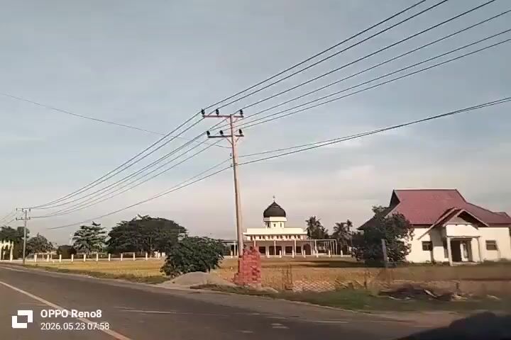 The video shows a landscape view from inside a moving vehicle. A road is visible on the left of the frame. On the right side, there is a grassy field with a mosque in the distance. The mosque has a dark dome and white walls. Overhead, there are power lines running across the sky. A black pickup truck with several people in the back drives past on the road. The sky is light blue with some clouds. In the lower left corner, there is a watermark indicating the video was taken on an OPPO Reno8 phone. The date and time are displayed as 2026.05.23 07:58.