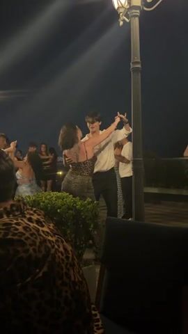 The video shows a group of people dancing at night outdoors. A woman with dark curly hair and a leopard print dress is dancing with a man wearing a white shirt and black pants. They are in the center of the frame, with other people dancing around them. In the foreground, a person wearing a leopard print shirt is visible from the chest up. A lamppost with a light fixture is in the upper right corner of the frame, casting light on the scene. There is a low wall or railing behind the dancers, and a dark sky is above. The environment appears to be an outdoor venue, possibly a restaurant or party, with some greenery visible in front of the dancers. The lighting is dim, with the lamppost being the primary light source.
