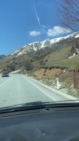 The video shows a car driving on a gray asphalt road on a sunny day. The sky is blue with a few scattered white clouds.  The road curves gently to the left. On either side of the road is vegetation, mostly green with some bare trees and brown patches.  In the background is a mountain range, parts of which are covered in snow. There are a few small structures visible on the lower slopes of the mountains. A guardrail runs along the side of the road.  A black car passes in the opposite direction at the start of the video.  A downed telephone pole is visible near the side of the road later in the video.  The view is from the interior of the car, looking out through the windshield.