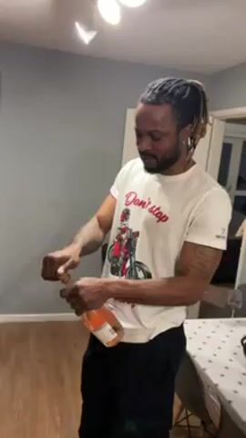 A man with dreadlocks, wearing a white t-shirt with a graphic and black pants, is standing in a room and opening a bottle of pink champagne. He removes the foil, pops the cork with force, and champagne sprays up into the air. The room has light gray walls, a white decorative table, and a dining area with chairs visible in the background.