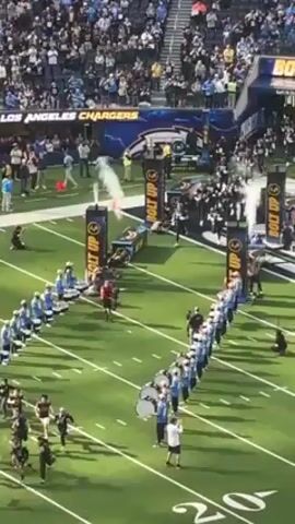 The video shows the Los Angeles Chargers NFL team running onto the field through a tunnel. There are four stands flanking the tunnel that say "Bolt Up" on the side and emit plumes of white smoke into the air. A marching band in light blue and white uniforms is lined up on either side of the tunnel. The stands are full of spectators. The football field is green and marked with white lines. A coach wearing a white shirt stands next to the band. A few cameramen kneel to capture the scene.