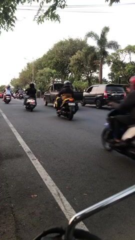 The video shows a street scene with traffic. Several motorcycles are visible, with riders wearing helmets. Some motorcycles are moving away from the camera, while others are approaching. A black SUV is also present on the road, as is a white truck. Trees and palm trees line the side of the road, and there are some decorative barriers. The sky is overcast. The video appears to have been filmed from a moving vehicle, possibly a motorcycle, given the close-up view of handlebars at the bottom of the frame. The overall impression is that of a normal street scene in a city or suburban area.