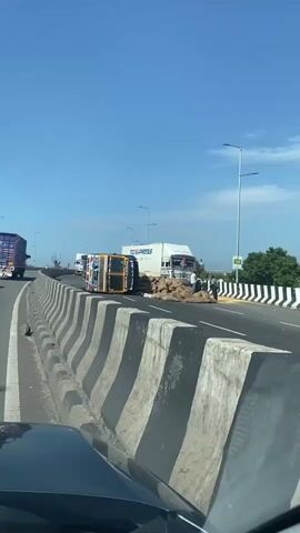 A flipped over truck lies on its side on a highway. Several sacks of cargo are spilled around the truck. A large white truck with the logo TCLEXPRESS is parked behind the flipped truck. Several people are standing around the white truck, some appearing to be examining the cargo. The sky is clear and blue.
