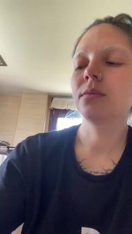 A person with dyed hair, a nose ring, and a tattoo on their chest takes a drink from a blue glass filled with a green liquid. They are wearing a black t-shirt and are in a room with light-colored walls. A window and some tiled walls are visible in the background.