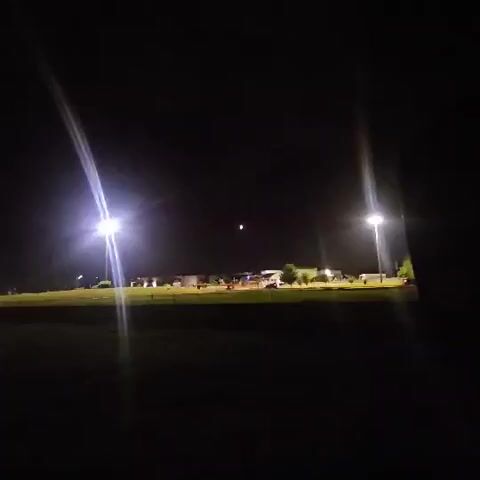 The video begins by showing a dark scene with artificial lights illuminating a wide, grassy field. In the distance, there are buildings and trees visible under the night sky. The camera then slowly pans to the right and zooms in slightly. A crescent moon is visible in the sky, appearing as a thin sliver of light. Artificial lights from what appear to be stadium lights or large outdoor lamps are prominent in the foreground, creating bright beams that extend across the frame. As the video progresses, the camera continues to focus on the moon, which becomes the central subject of the frame. The surrounding buildings and landscape recede into darkness. The video ends with a close-up view of the crescent moon against the black night sky.