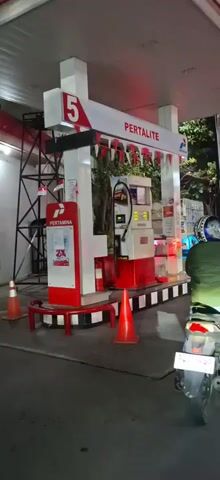 The video shows a gas station at night. A Pertamina gas pump with the label PERTALITE is visible, with the number 5 on top. There are several orange traffic cones around the pump. A person on a motorcycle is waiting to refuel, with the back of their green jacket and helmet visible. Further down the road, there is a line of other motorcycles and a white car, indicating a queue at the gas station. The scene is lit by the gas station's lights and the headlights of the vehicles. The atmosphere appears to be busy.